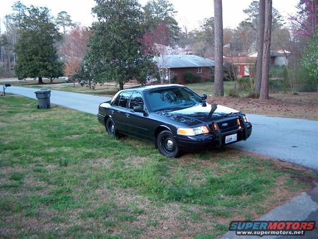 Black 2001 Ford Crown Victoria Police Interceptor parked on a grassy shoulder next to a road. It has a push bar and light bar