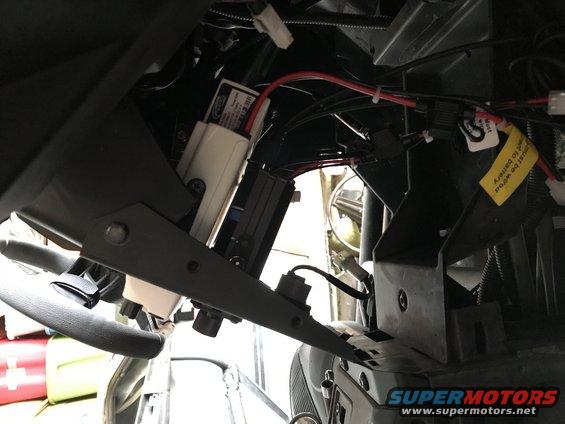Interior view of vehicle wiring harness with a white box and red wires, yellow sticker reads "must be wired to battery"