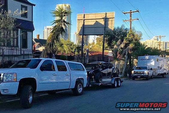 White pickup truck towing a trailer with a side-by-side, and another pickup towing a travel trailer