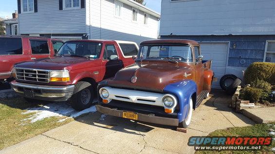 1950s Ford pickup truck with blue fender, 1990s Ford Bronco, and red SUV in driveway