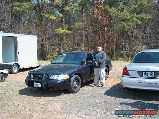 Dark blue Ford Crown Victoria with a push bar and a man standing next to it. Silver Mercury Marauder parked to the right. White trailer in background
