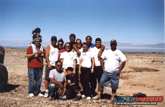 Group of people posing in a desert with a body of water in the background. One person is on another's shoulders