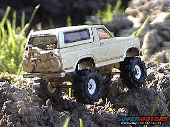 Toy 1979 Ford Bronco with large off-road tires and a motorcycle on the rear rack, sitting on dirt and grass