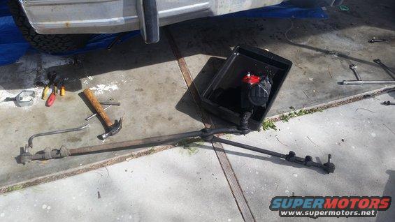 Steering linkage and tools laid out on concrete, with part of a vehicle in the background