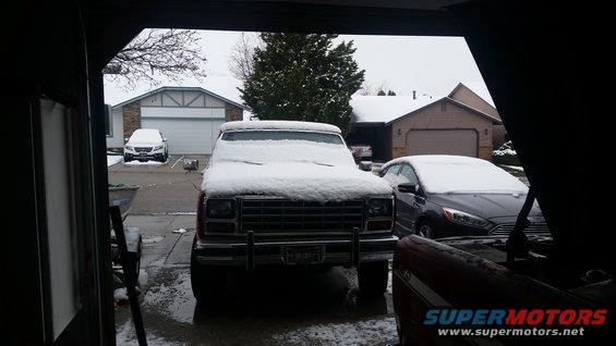 Front view of an old SUV, possibly a Ford Bronco, covered in snow. Another car is parked to the right, also snow-covered. Houses and a tree are in the background