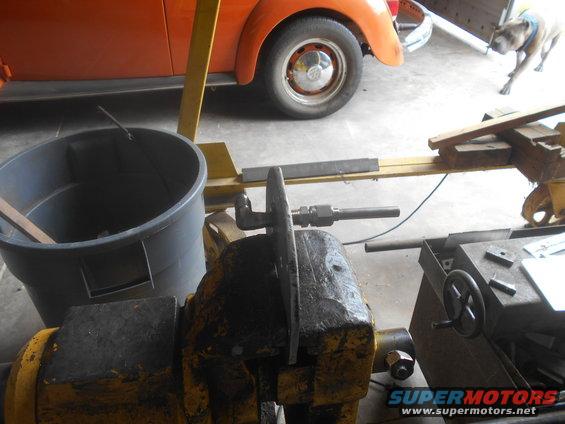 Metal part clamped in a yellow vise, with an orange car and a dog visible in the background