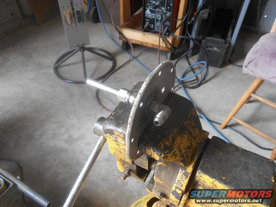 metal fuel sending unit assembly clamped in a yellow vise, with workshop equipment in background