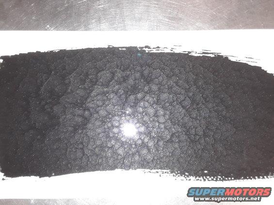 Pile of black granular material on white paper, with a metallic surface above