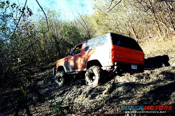 Orange Ford Bronco with lifted suspension and large tires driving up a muddy hill in a wooded area