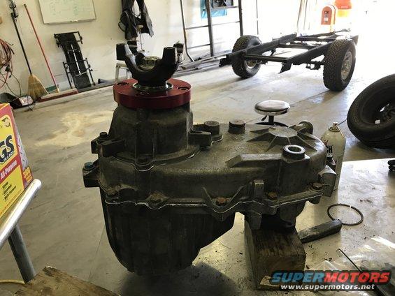 Transfer case with red adapter plate and U-joint, resting on wooden blocks. Vehicle chassis in background