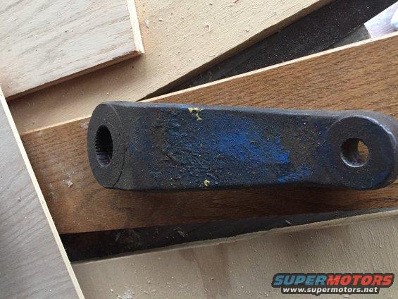 Black metal automotive steering linkage part with blue paint markings, resting on wooden planks