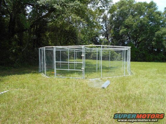 Octagonal wire mesh enclosure with metal frame and double doors, set in a grassy field with trees in the background. A roll of wire mesh lies on the grass