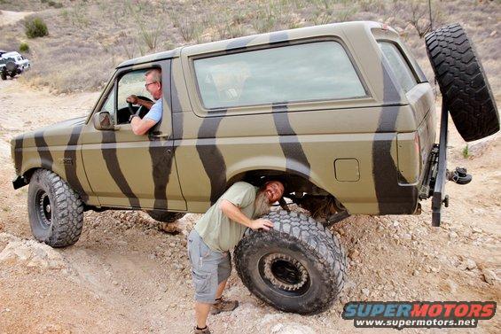 1996 Ford Bronco with camouflage paint off-roading, one person driving, another under the vehicle checking a tire