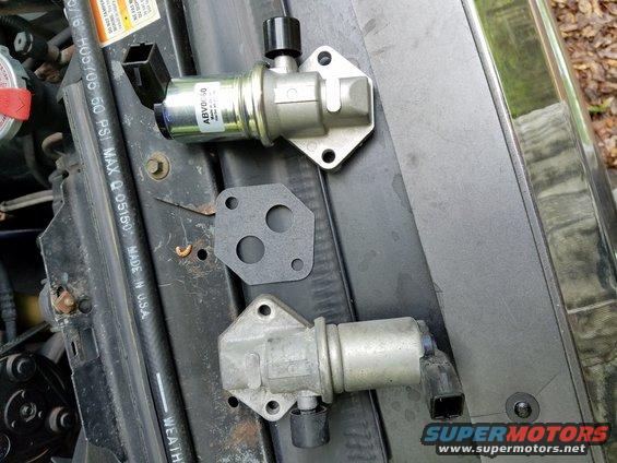Idle air control valve and gasket on engine bay surface. One valve is labeled ABVD60
