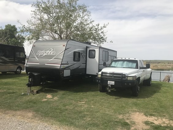 White pickup truck with a heavy-duty bumper towing a Pioneer travel trailer. Another RV is parked to the left