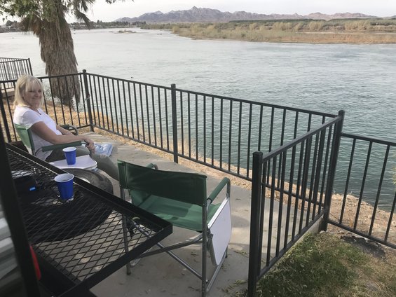 Woman in white shirt sits in green chair on concrete patio overlooking river, with mountains in distance