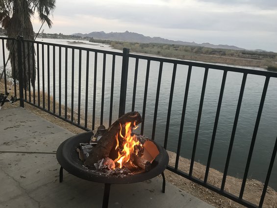 Fire pit with burning logs on a concrete patio, overlooking a wide river and distant mountains under a cloudy sky
