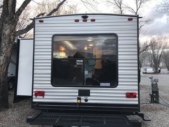 Back of a travel trailer with a large window showing the interior. A tree is to the left