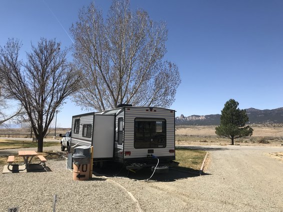 Travel trailer parked at campsite with pickup truck behind it. Picnic table nearby. Mountains in background