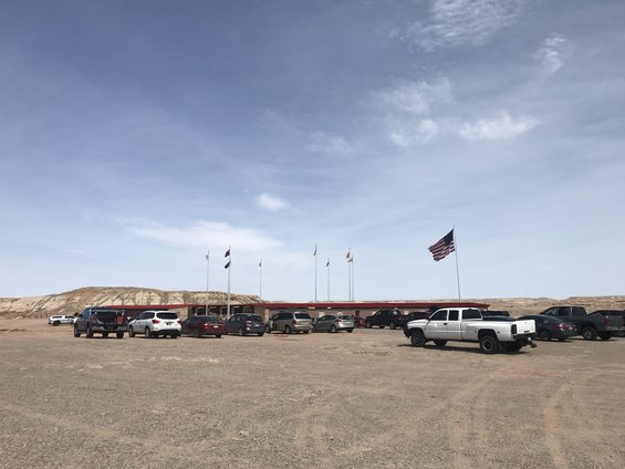 Parking lot with several cars and trucks in front of a building with flags, under a partly cloudy sky