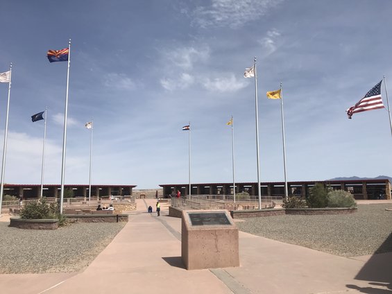 Flags fly in front of a long, covered building complex under a partly cloudy sky