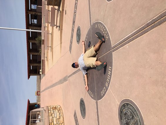 Person lying on the ground at Four Corners Monument, with text "IN FREEDOM UNDER GOD" visible