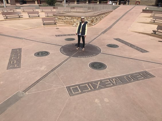 Woman in vest and pants stands at Four Corners Monument, with state names and seals visible on the ground