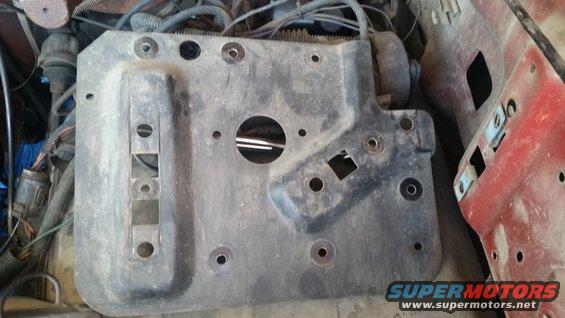 Gray metal engine bay bracket with multiple bolt holes and cutouts, next to red painted metal