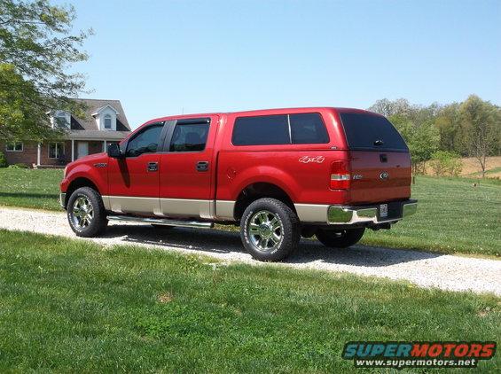 Red pickup truck with tan lower body, black camper shell, and chrome wheels parked on a gravel driveway