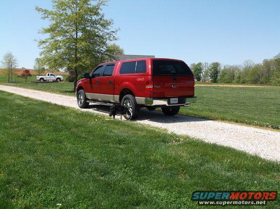 Red pickup truck with camper shell parked on gravel driveway. Another white pickup truck is visible in the background