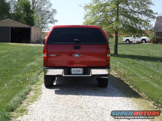Rear view of a red Ford F-150 pickup truck with a cap. Another white pickup truck is visible in the background