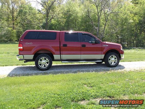 Side view of a red and tan Ford F-150 pickup truck with a camper shell parked on a gravel driveway
