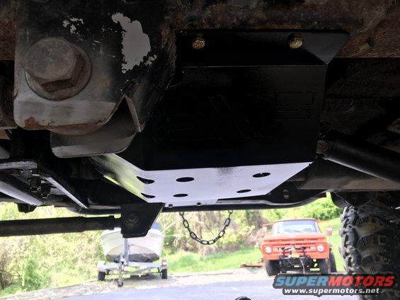 Underside view of a vehicle frame with a skid plate installed. An orange truck and a boat on a trailer are visible in the background