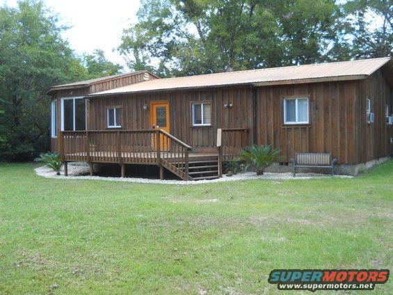 Wooden house with a deck and orange door, surrounded by grass and trees