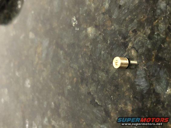 Close-up of a brass carb jet with a small pin extending from it, resting on a speckled surface. Supermotors.net logo visible