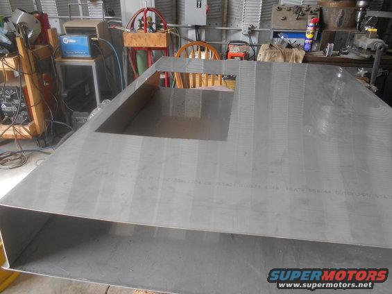 Metal sheet with a rectangular cutout, sitting on a workbench in a garage with tools and welding equipment in the background