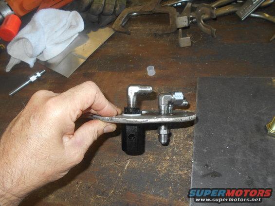 Hand holding a metal fuel pump assembly with two angled fittings and a black central component