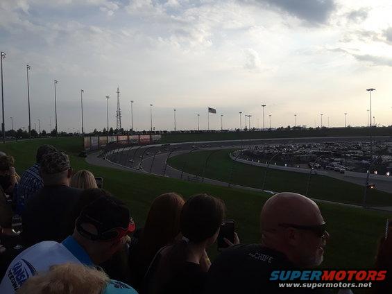 people watching a race track with lights and a parking lot full of cars