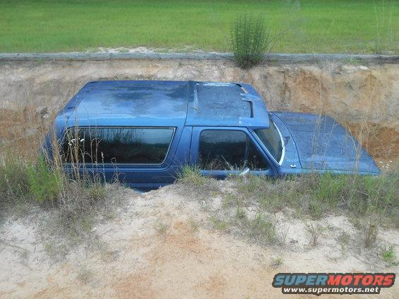 1992 Ford Bronco partially buried in a dirt ditch with grass growing around it