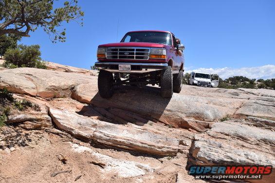 Red 1995 Ford Bronco front view, lifted, on rocky terrain with a white Jeep in background