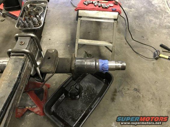 Truck axle spindle with leaf springs and U-bolts, resting on a red jack stand. A drip pan with oil is below the spindle. Tools and parts are on a metal stand in the background