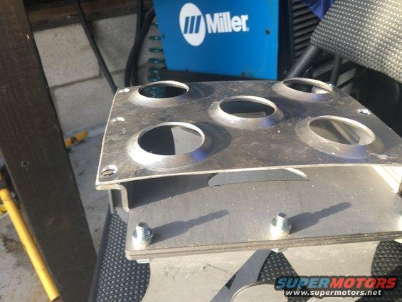 Metal plate with five circular holes, bolted to another metal piece. Miller welding machine in background