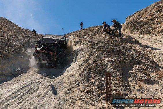 Modified off-road vehicle going down a steep sandy slope with people watching from above