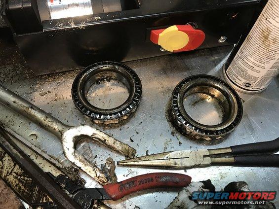 Two used tapered roller bearings on a metal workbench with pliers, a puller, and a can of spray