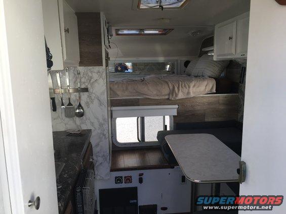 Camper van interior with bunk bed, kitchen, and table. Supermotors.net logo visible