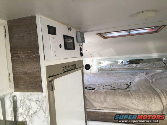 Camper interior showing a bed, refrigerator, and control panel