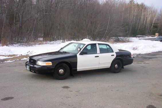 Black and white police car parked on asphalt with snow in background. Looks like a 2001 Ford Crown Victoria