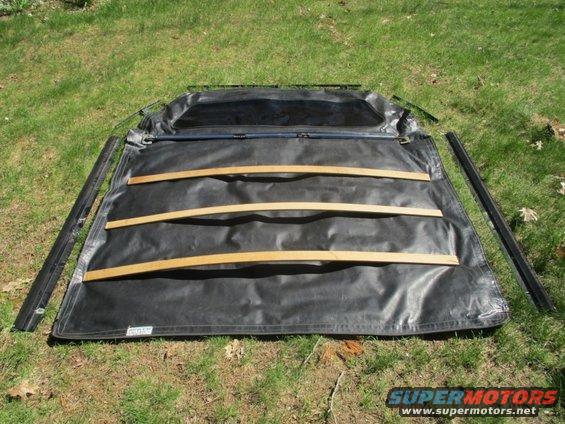 Unfolded black soft top with wooden support slats and metal rails, lying on grass