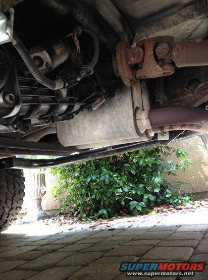Underside view of a vehicle's transmission, driveshaft, and exhaust system. A tire is visible on the left