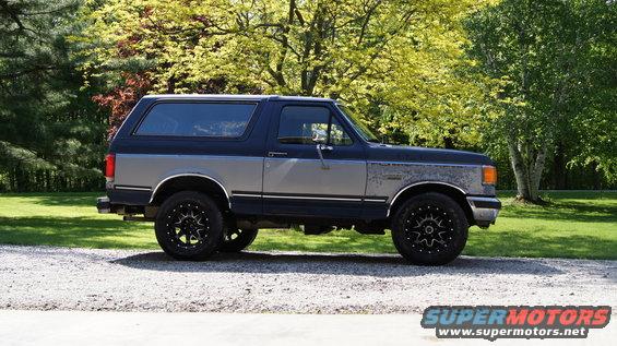 Two-tone blue and gray 1987 Ford Bronco with aftermarket black wheels parked on a gravel driveway